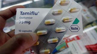 Close-up image of a hand holding a strip of Tamiflu pills in front of a packet of them. The pills, which are half yellow and half white, can be seen in the pill strip. The pill packet behind is white and has branding on it. 